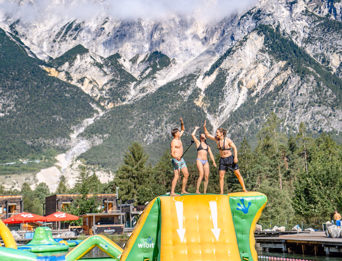 drei Personen stehen auf einer aufblasbaren Rutsche und High-Fiven. Sie sind auf einem See vor einem Gebirgspanorma